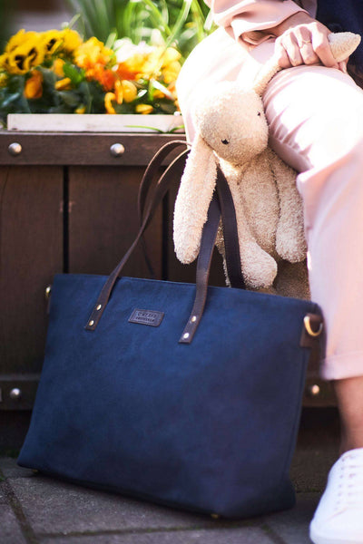 Navy Wax and Dark Brown Diaper bag by O My Bag on the ground outside next to parent with stuffed animal.